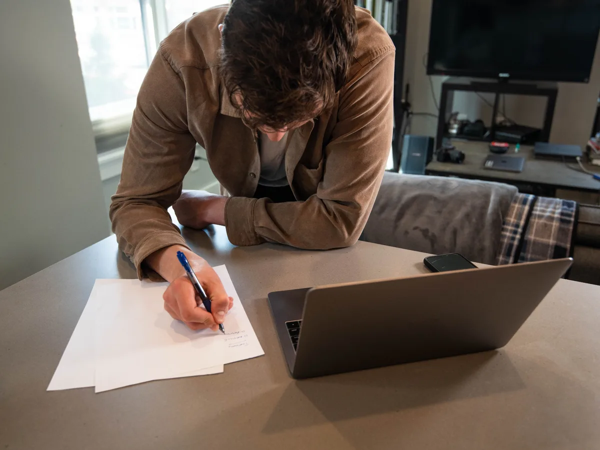 A man writing figures on paper beside his laptop at a kitchen table