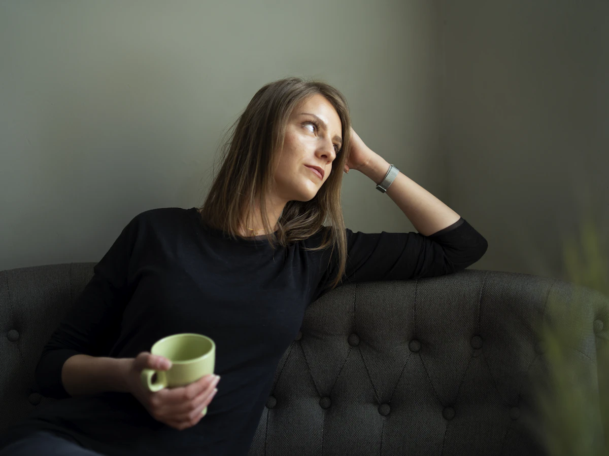 A woman sitting calmly on a couch with a mug, gazing toward a window