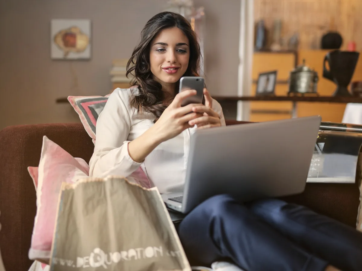 A woman on a couch with a shopping bag, checking her phone beside her laptop