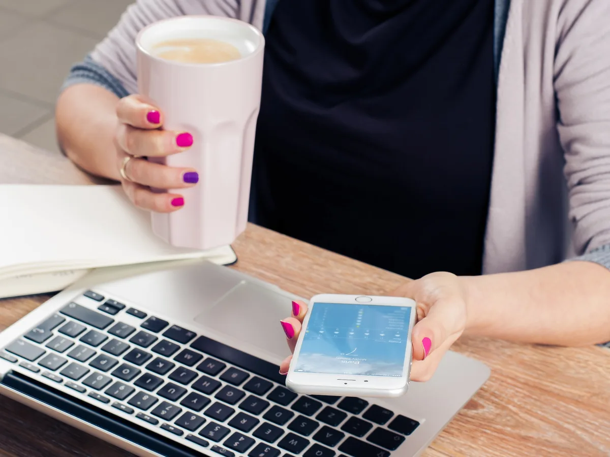 Hands holding a coffee and a phone over a laptop and an open notebook