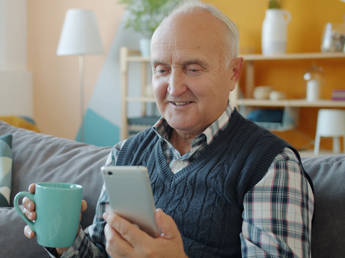 A smiling older man checking his phone on the couch with a mug of coffee